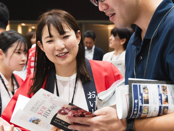 赤いハッピを着た女性がパフレットを読む男性に話しかけている写真